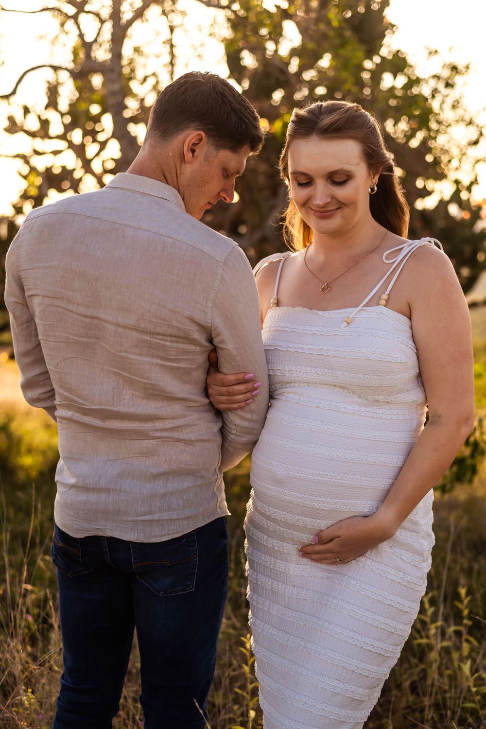 Expectant couple sharing an intimate moment in a golden sunset field while wearing coordinated casual attire.