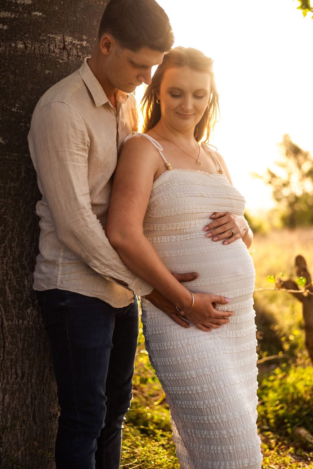 Couple poses against tree during golden hour maternity session in nature.
