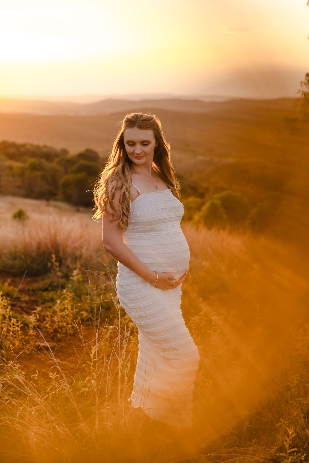 Maternity portrait of expectant mother in white dress standing in golden sunlit field at sunset.
