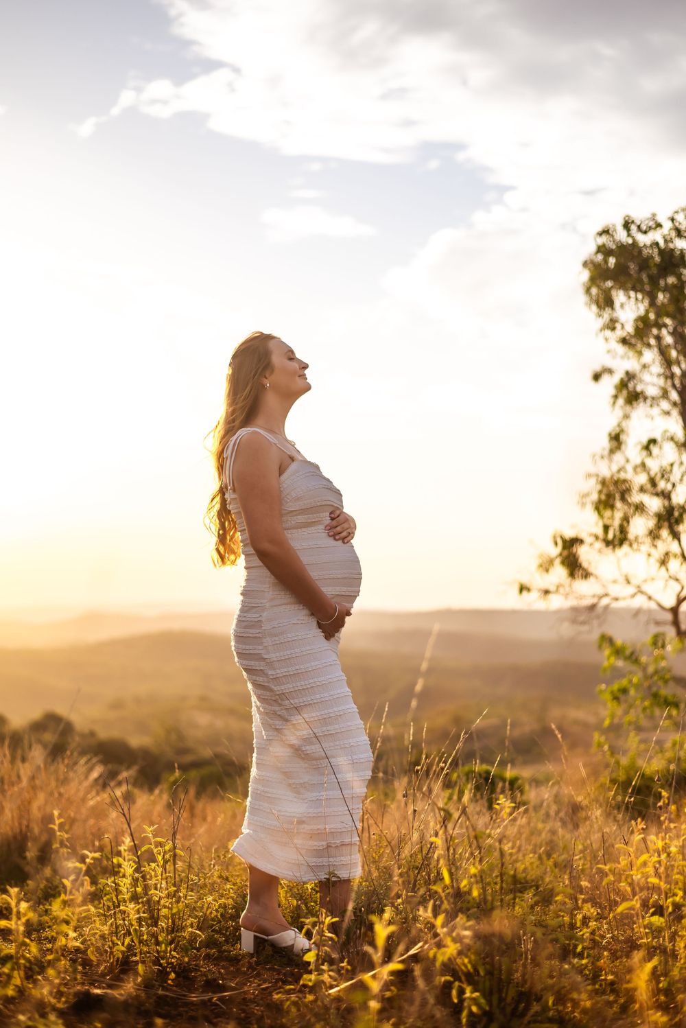 Pregnant woman in white dress poses against scenic sunset landscape with mountains in background.
