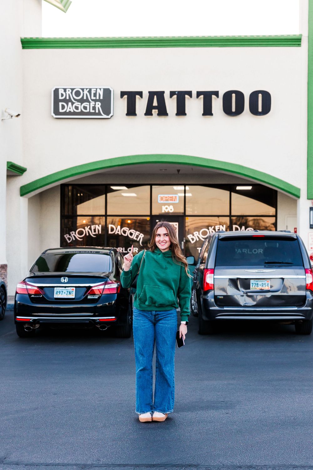Person standing in front of Broken Dagger Tattoo shop with parked cars.