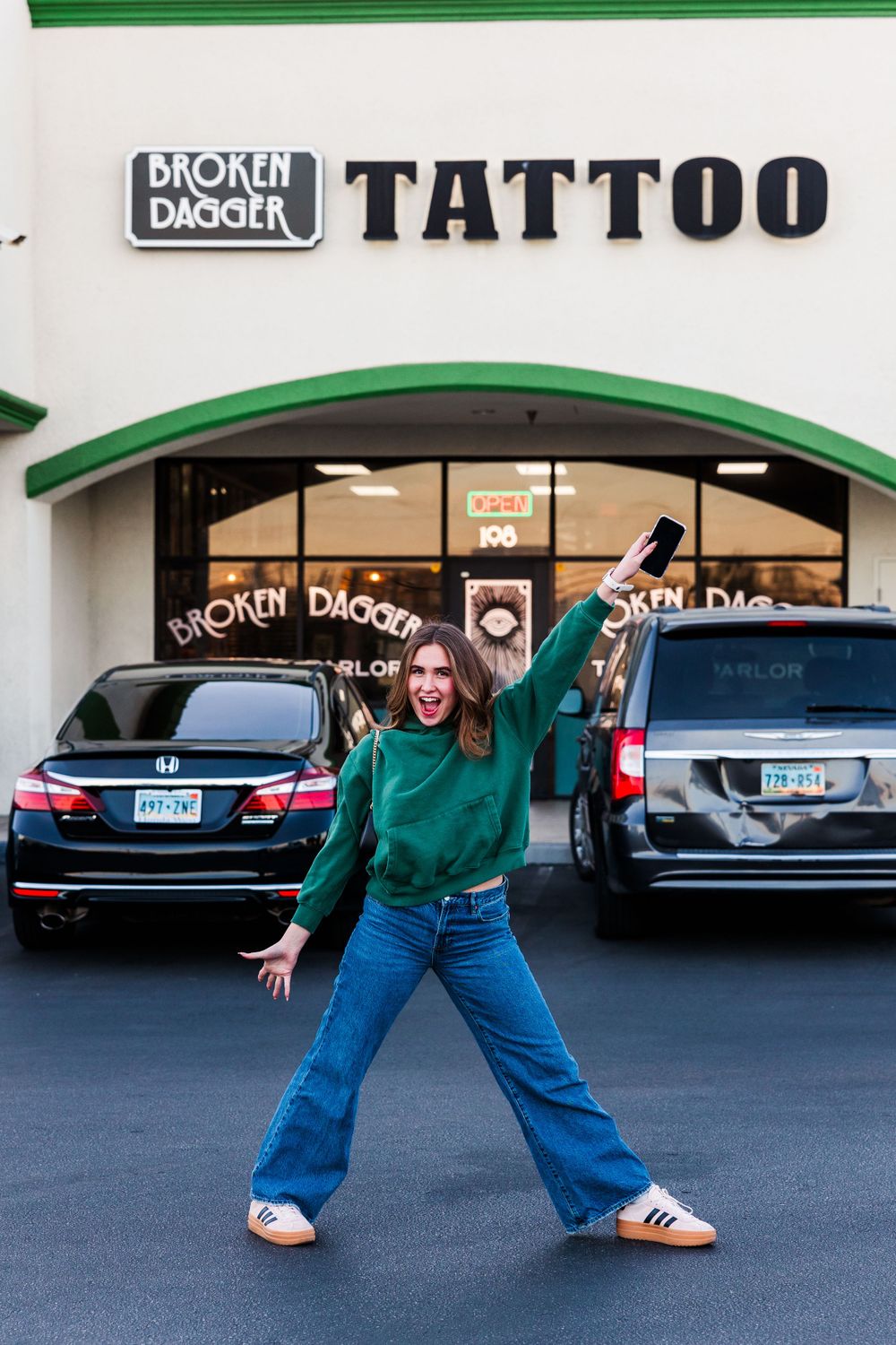 Energetic pose outside Broken Dagger Tattoo shop wearing green sweater and jeans.
