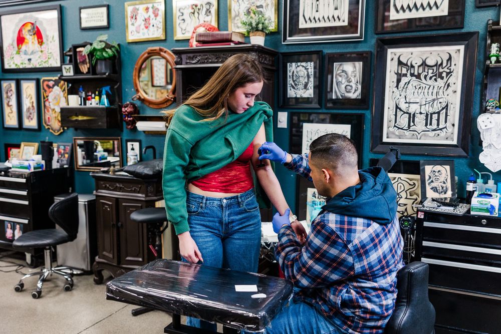 Tattoo artist in plaid shirt prepares their station while consulting with client in a studio decorated with art on walls.