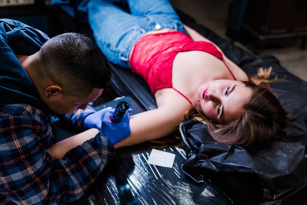 Tattoo artist in plaid shirt concentrates while working on reclining client.