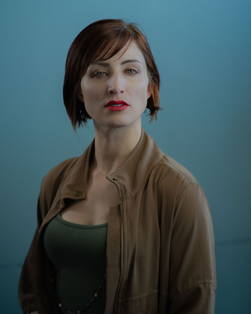 Dramatic portrait against teal background wearing brown jacket.