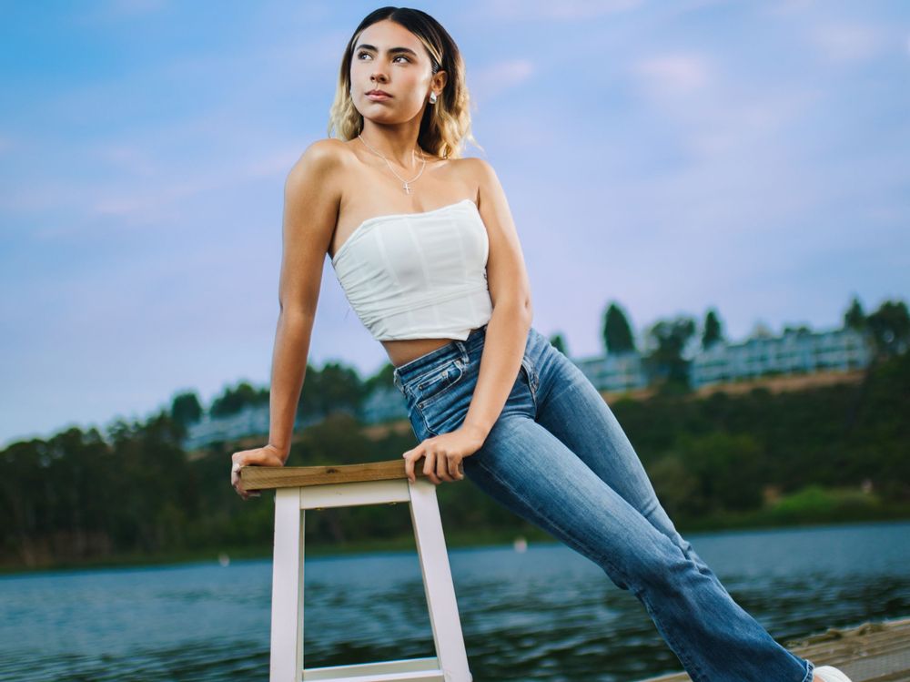 Casual portrait on white stool by lake wearing white top and jeans.