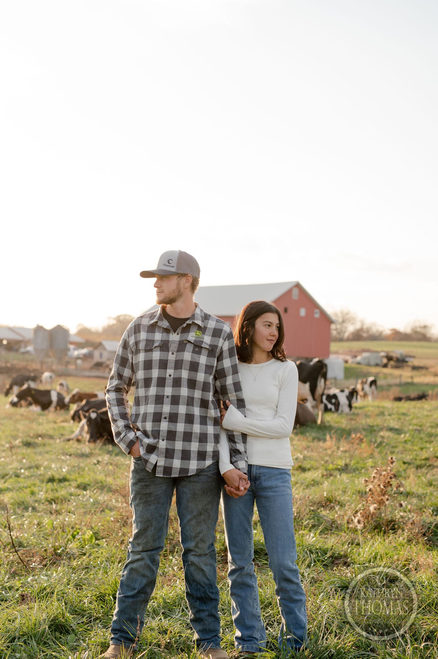 A couple stands together in casual attire on their farmland with cattle grazing behind them.