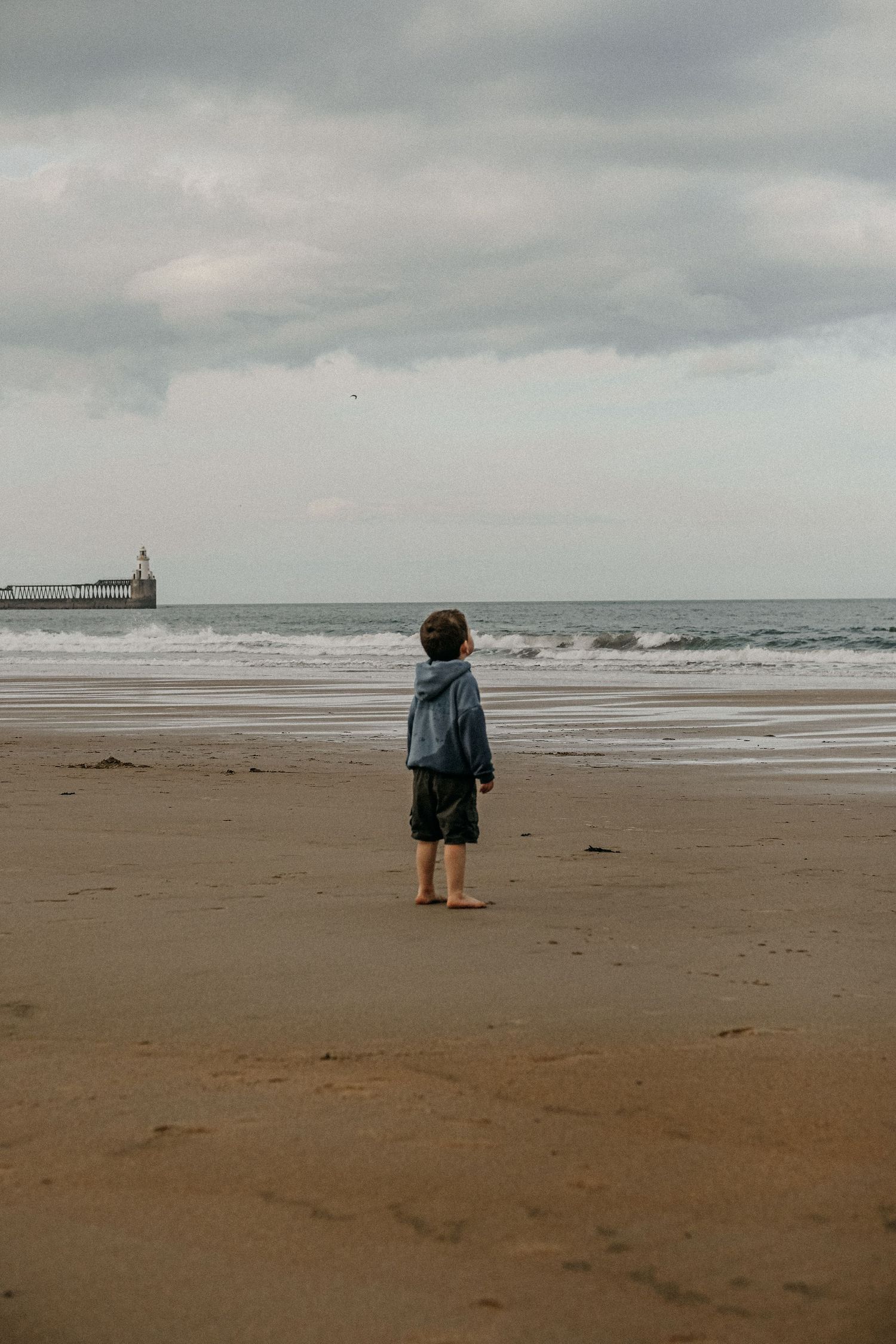 Small child stands alone on a moody beach watching waves with a pier in the distance.