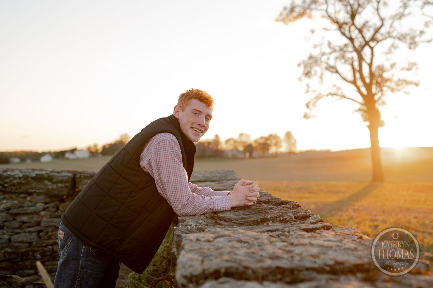 Person in vest smiling while leaning on stone wall during golden hour.