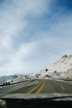 Snow-covered mountain road curves through winter landscape under cloudy blue skies.