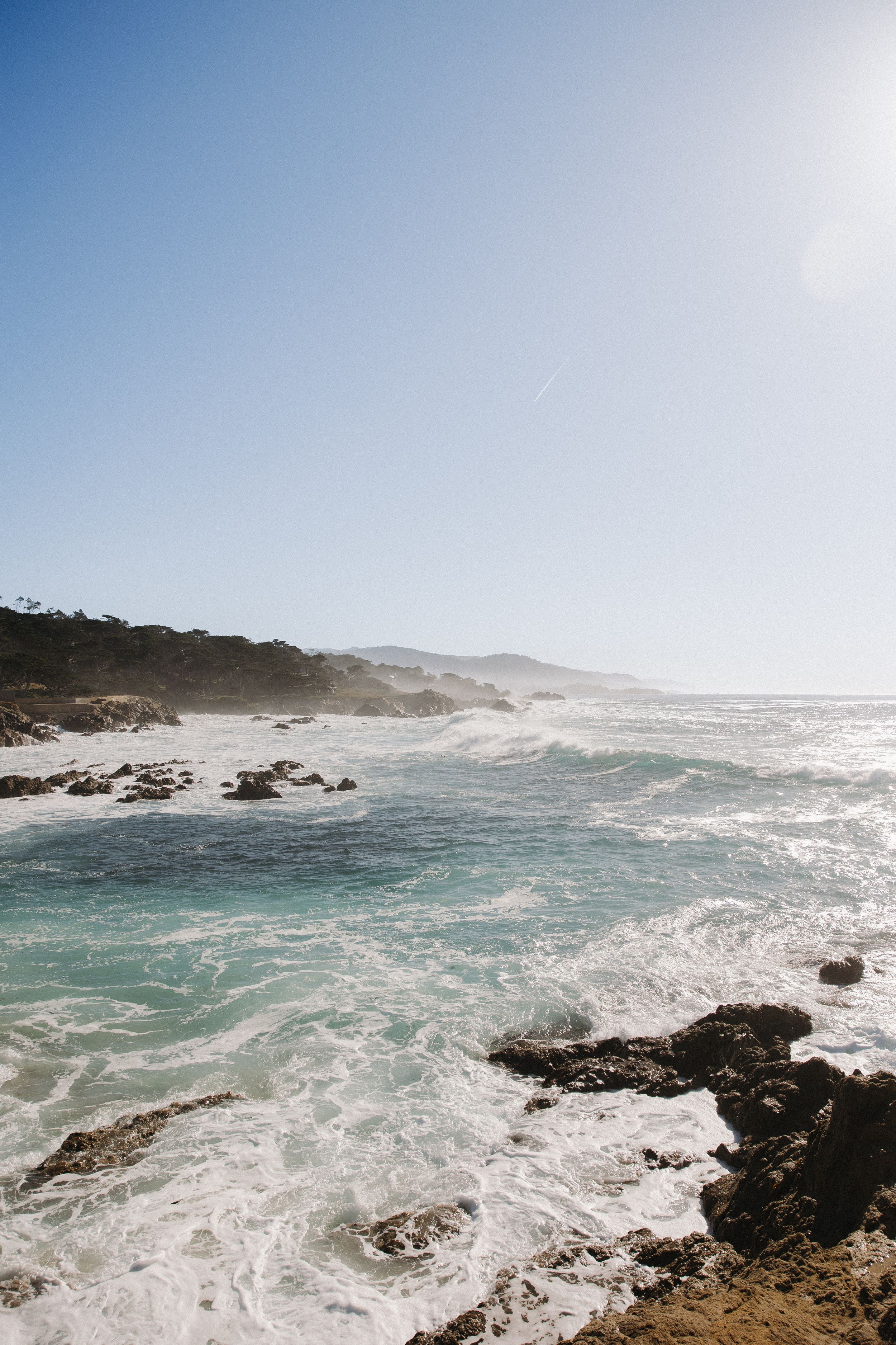 Dramatic coastline with turquoise waves crashing against rocky shore under bright blue sky, Carmel By The Sea, California.