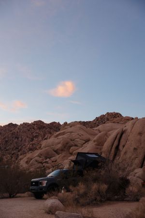 A vehicle with rooftop camping setup parked among large desert rock formations during sunset at Indian Cove in Joshua Tree.