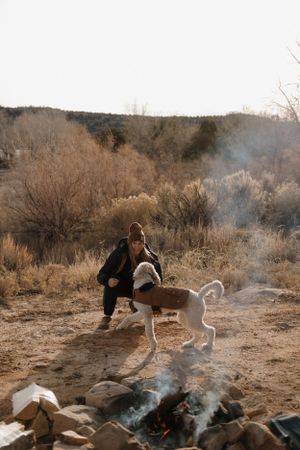 A dog plays near a smoking campfire in a desert landscape with brush and mountains in the background.
