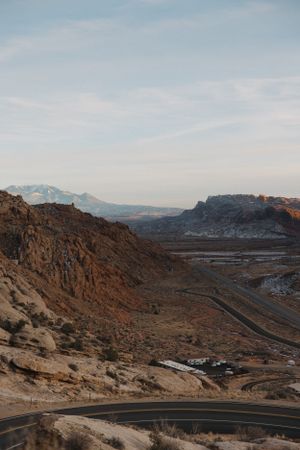 Winding mountain road cuts through dramatic red rock desert landscape under blue sky, Arches National Park.