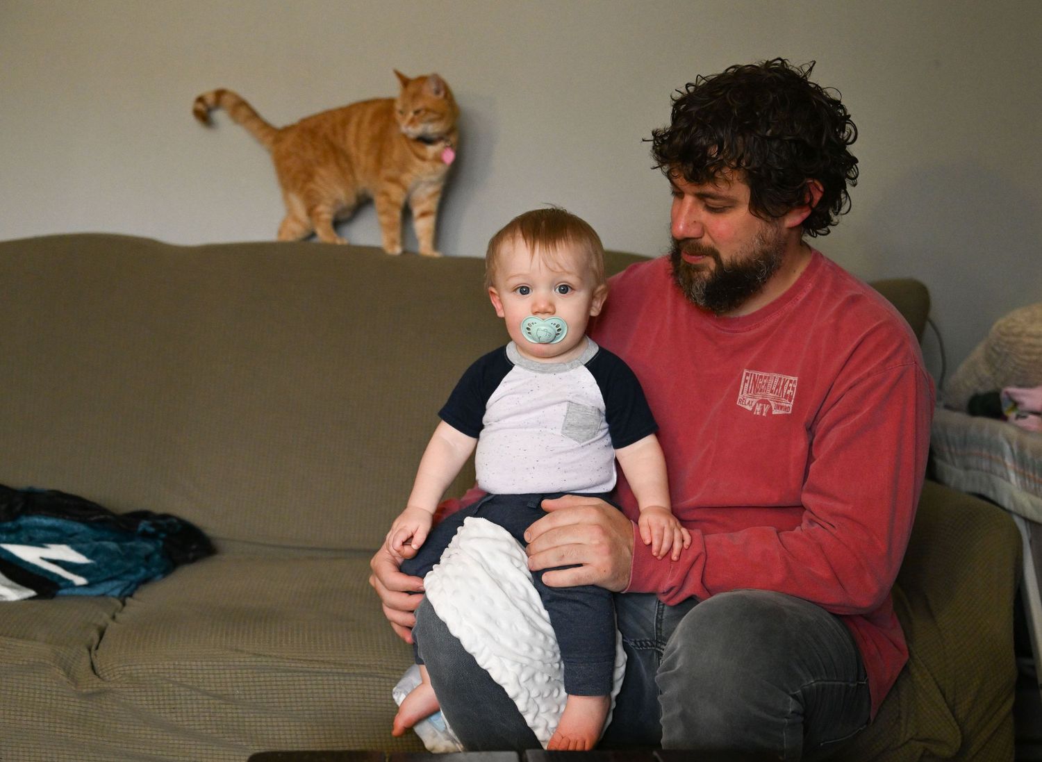 A photo of a man sitting on a couch holding his toddler in his lap with an orange cat on the back of the couch behind them