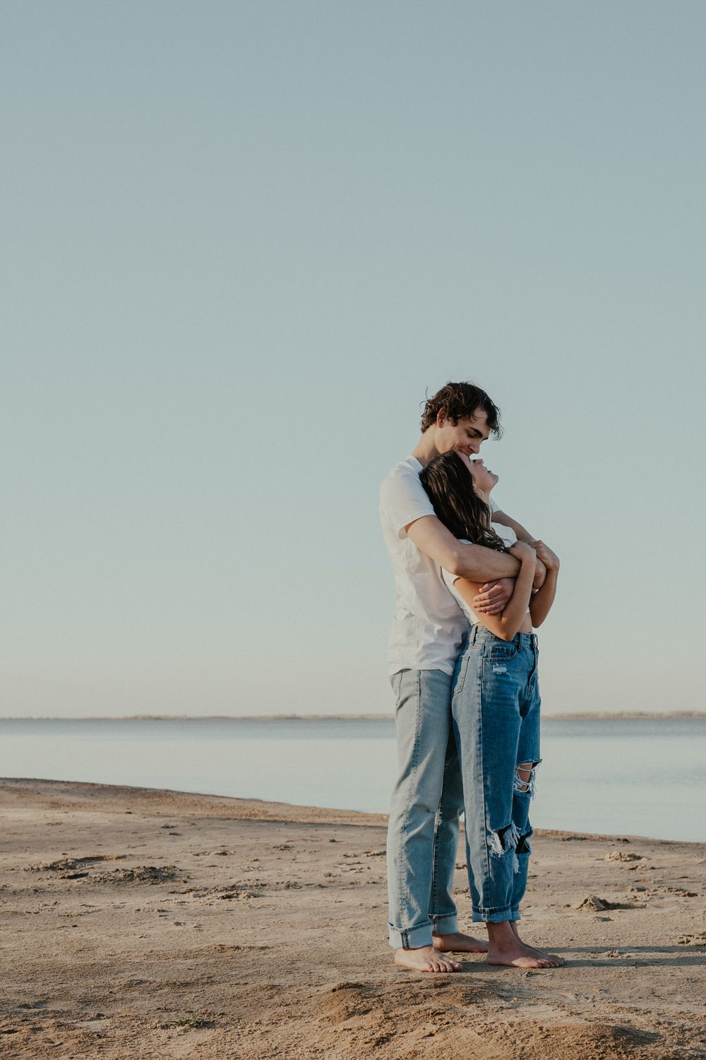 Couple in casual wear standing close together on beach at sunset.
