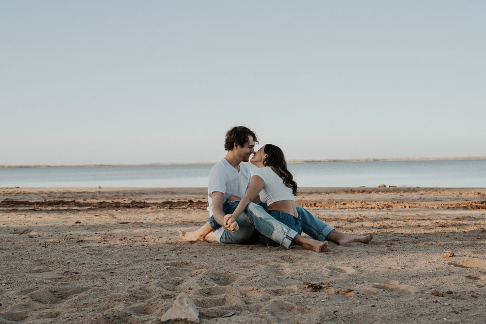 Couple in casual attire sitting close together on sandy beach near water.