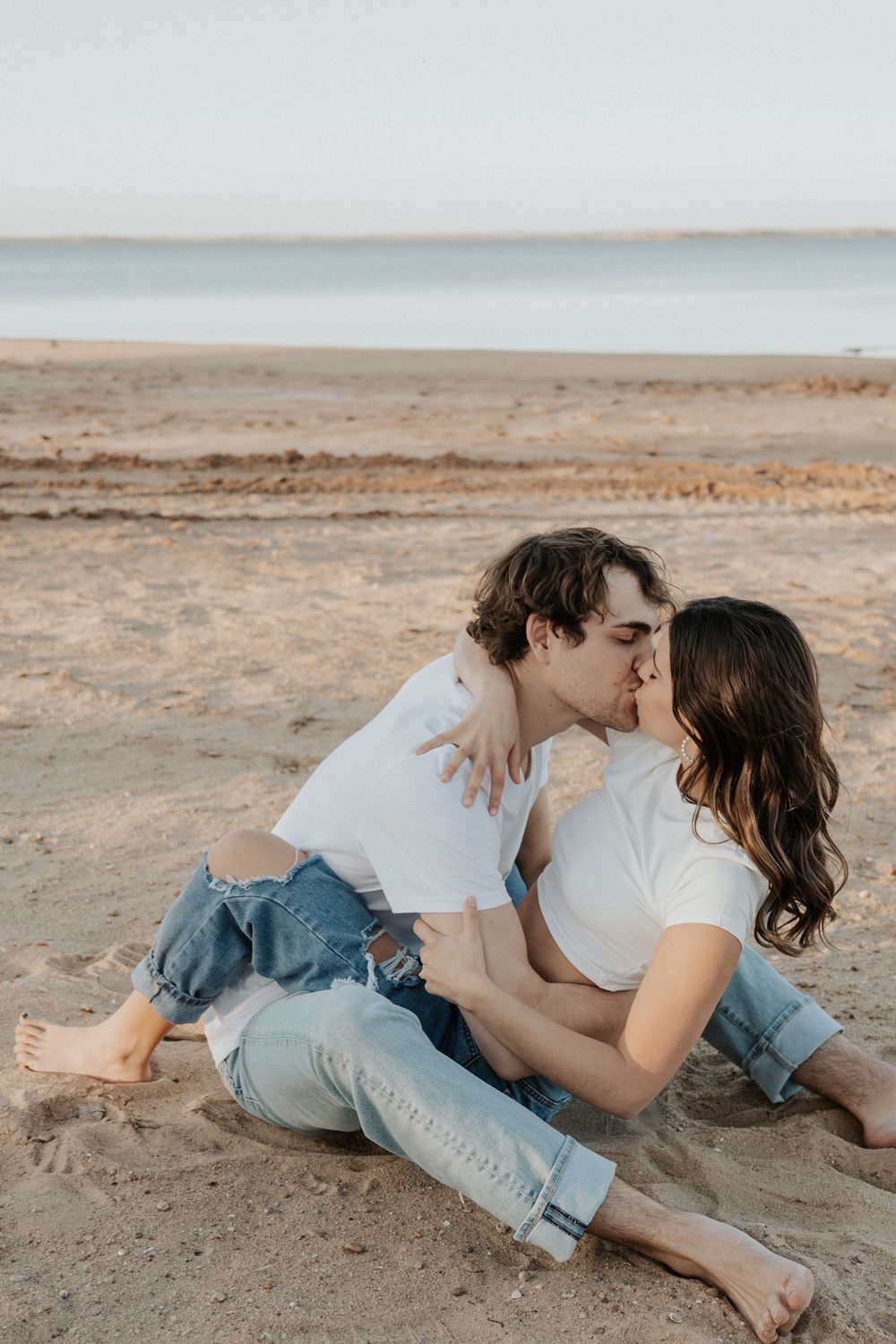 A romantic couple in casual white shirts and jeans share an intimate moment on a sandy beach at sunset.
