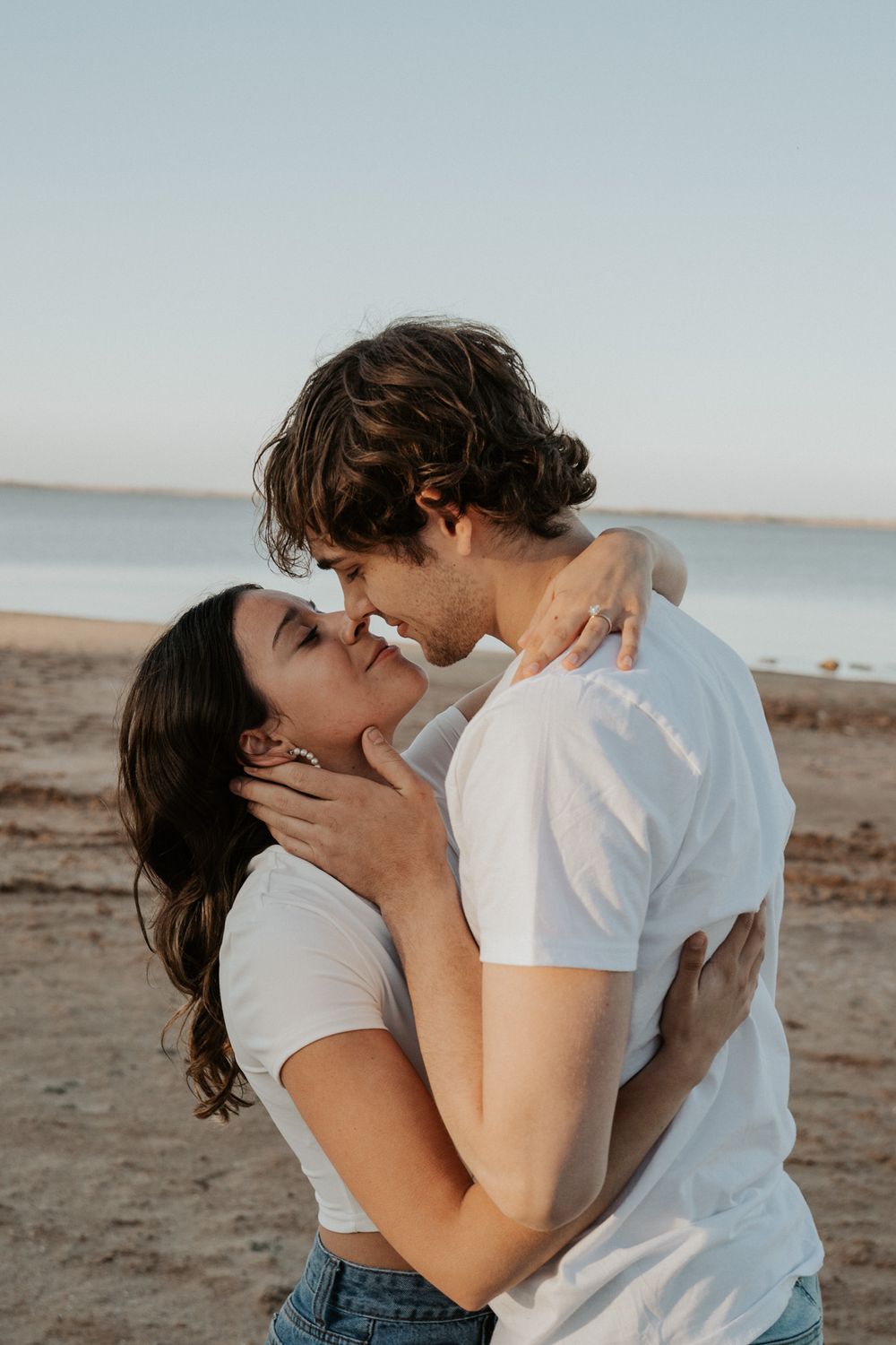 A romantic couple in white t-shirts embrace tenderly on a beach at sunset.