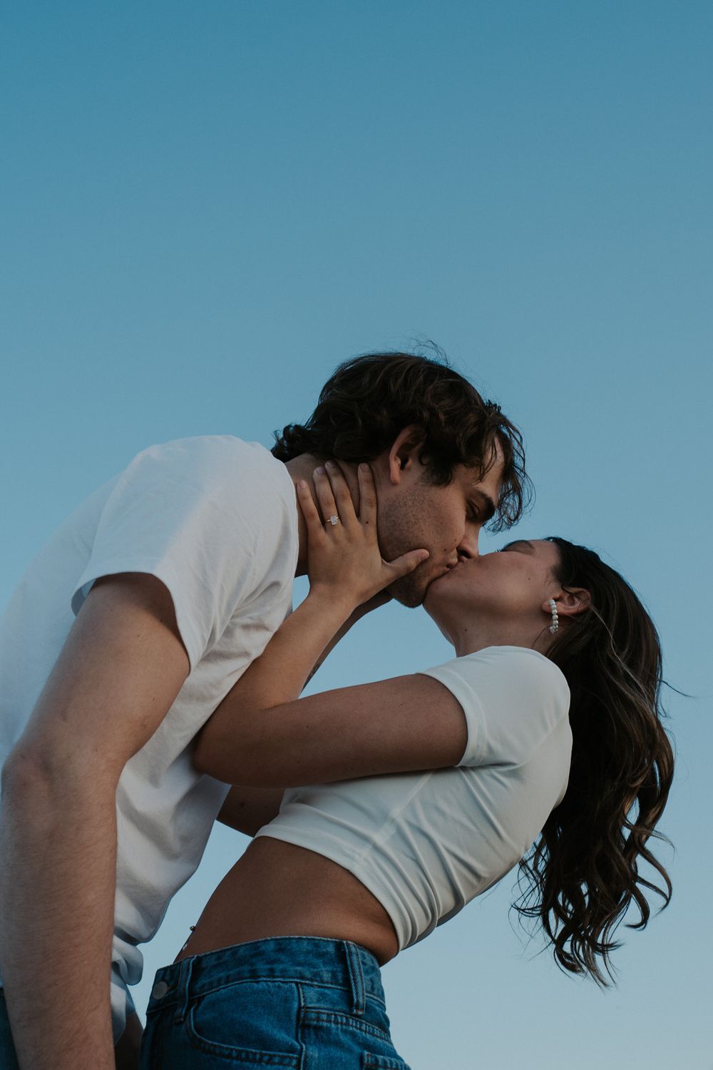 Intimate moment between couple in white shirts against blue sky background.