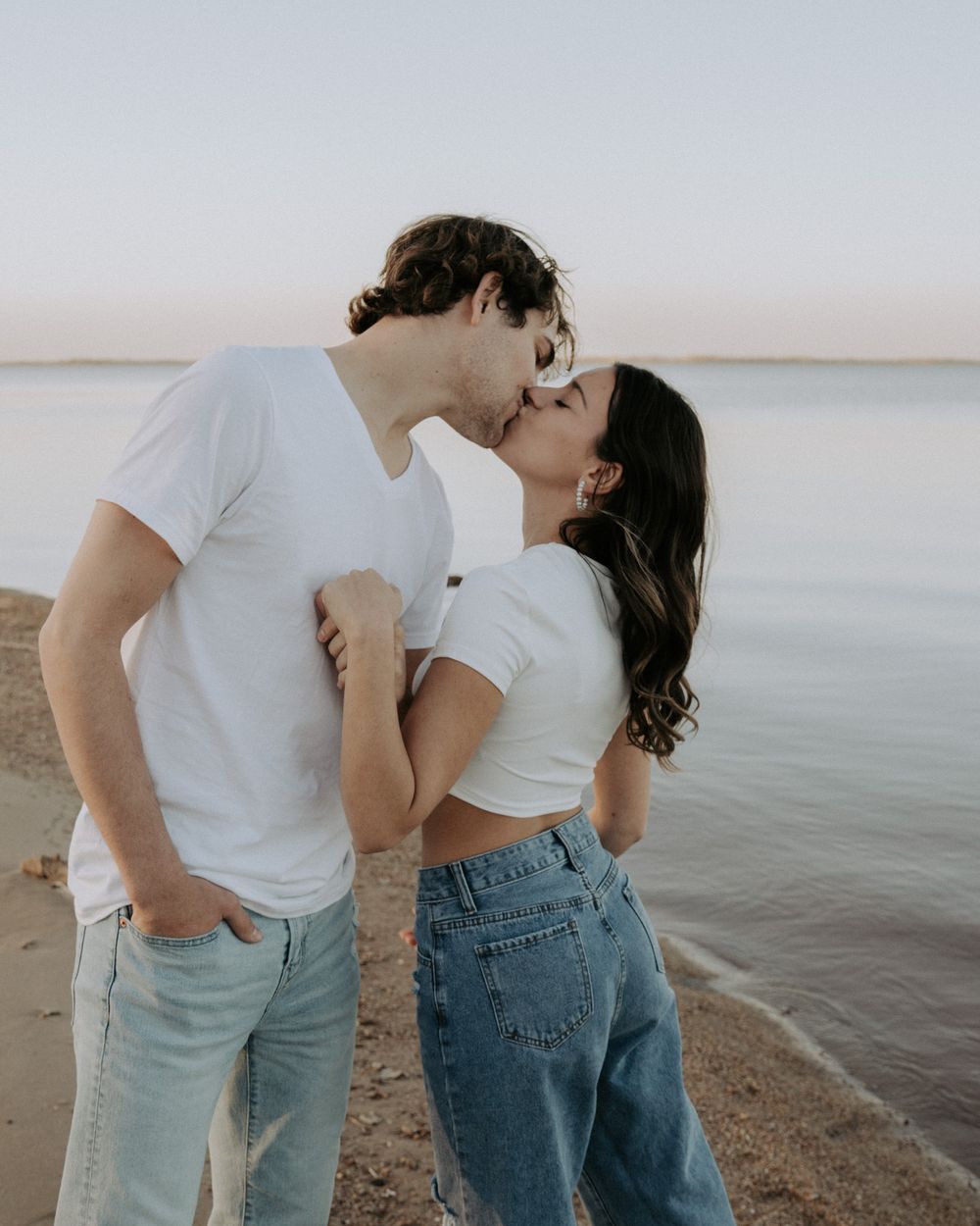 Couple in coordinated white tops and denim embrace on a peaceful beach.