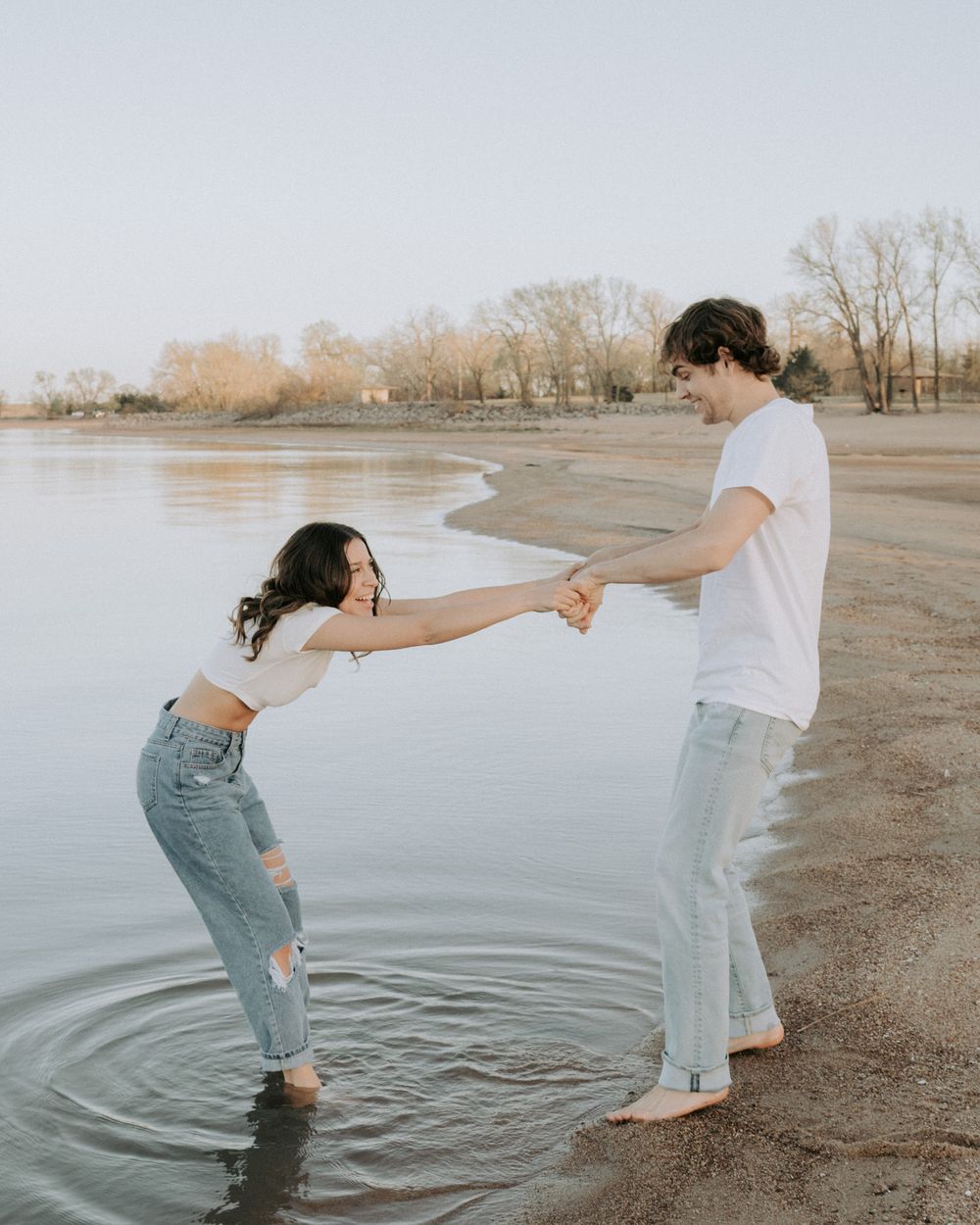 Playful couple holding hands and splashing in the water at a lakeside beach.