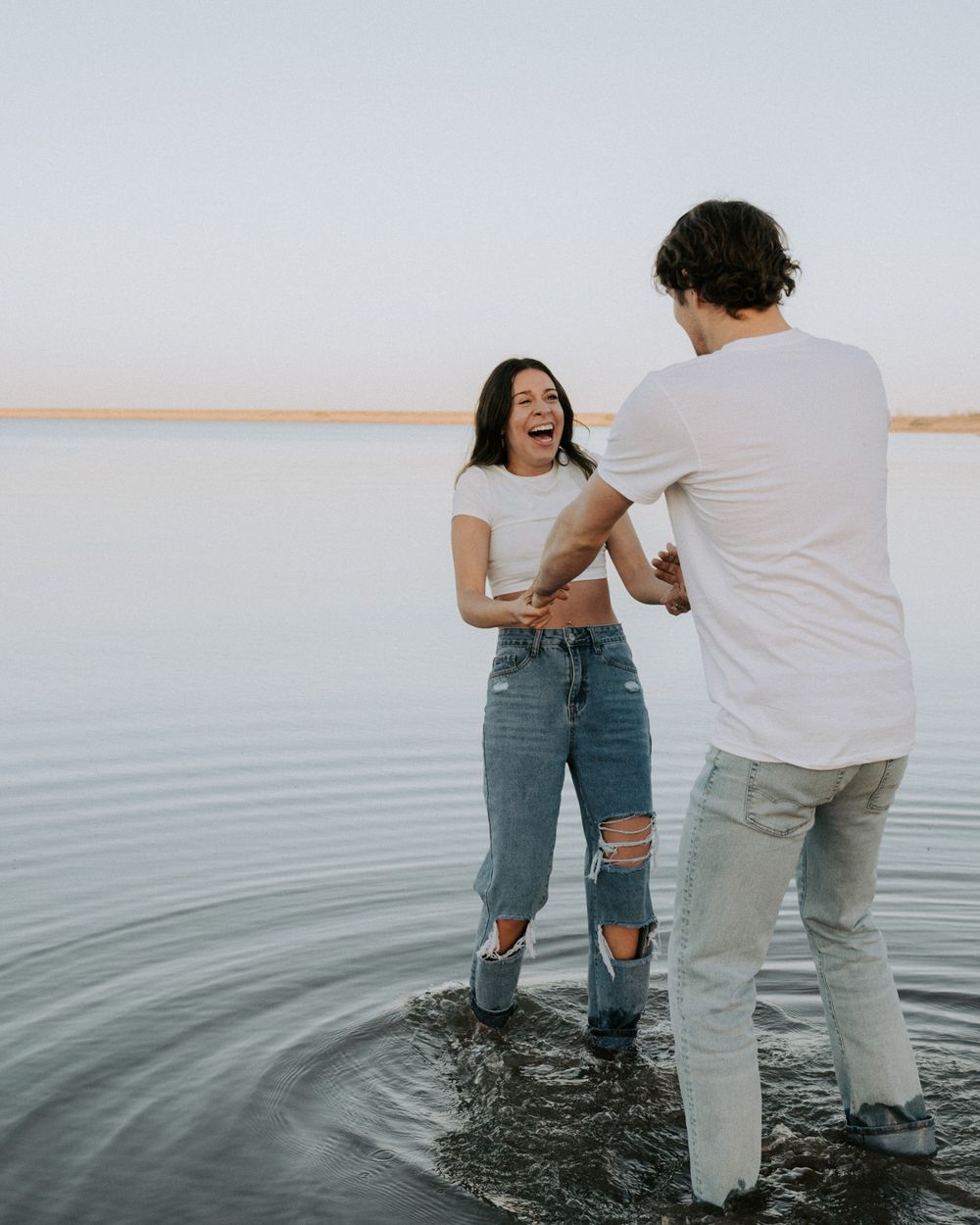 Couple playing and laughing together while standing in shallow water at sunset.