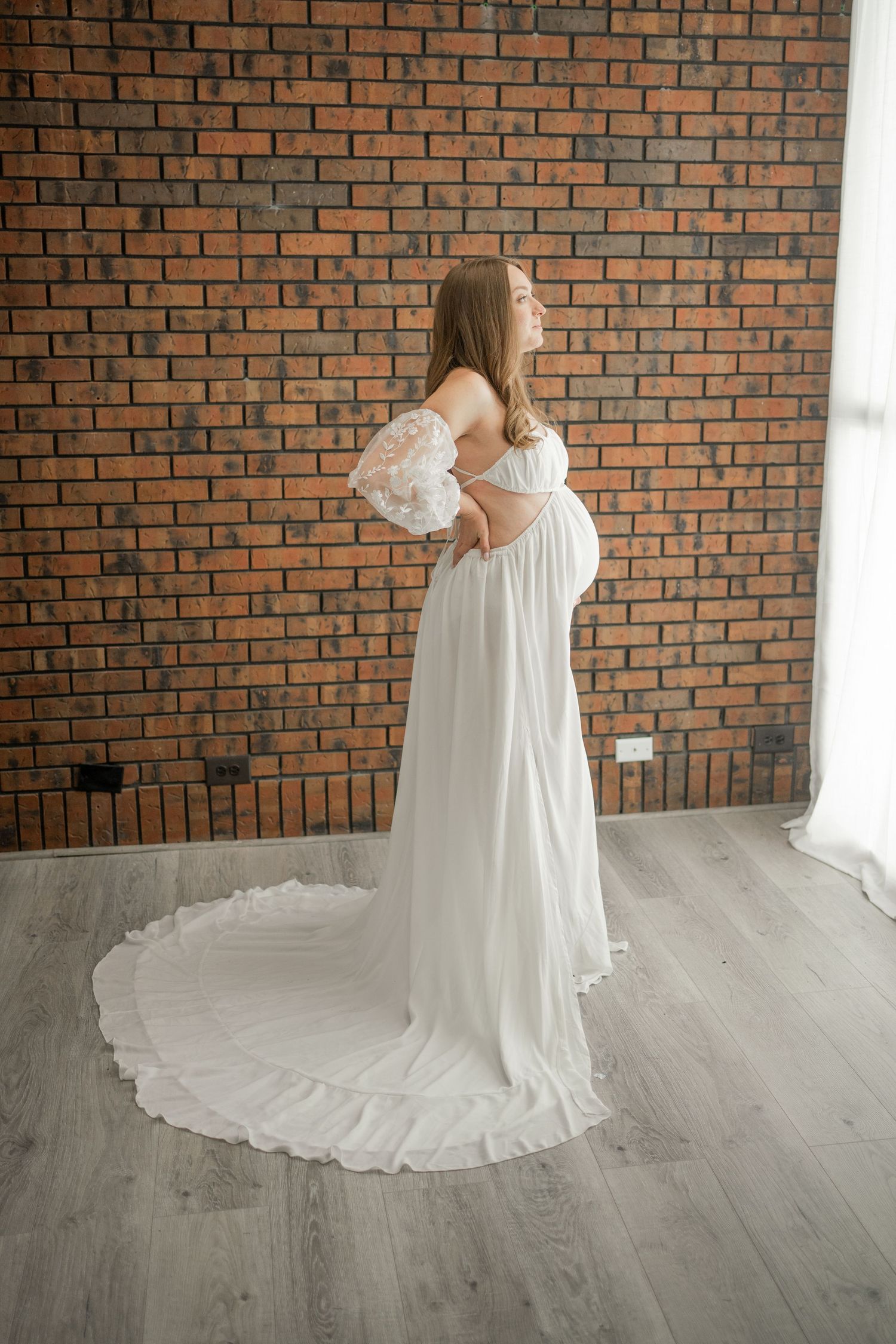 Expectant mother in flowing white dress poses against brick wall.