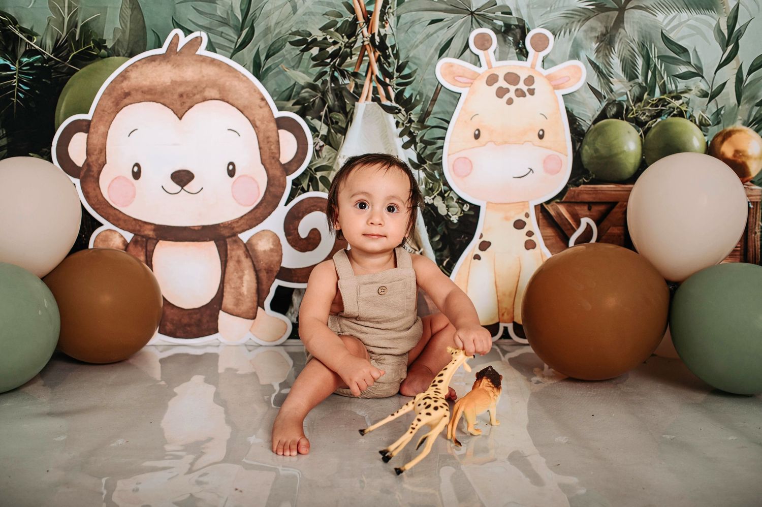 Safari-themed baby photo setup with monkey and giraffe decorations, balloons in brown and sage green colors, and toy giraffe.