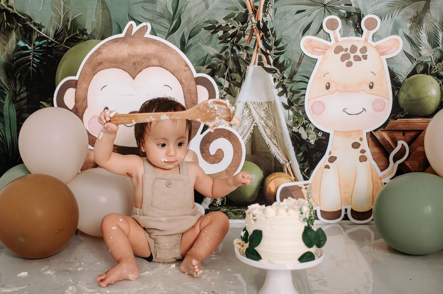 Jungle-themed photo setup with cartoon monkey and giraffe decor, white cake display, and balloon arrangement.