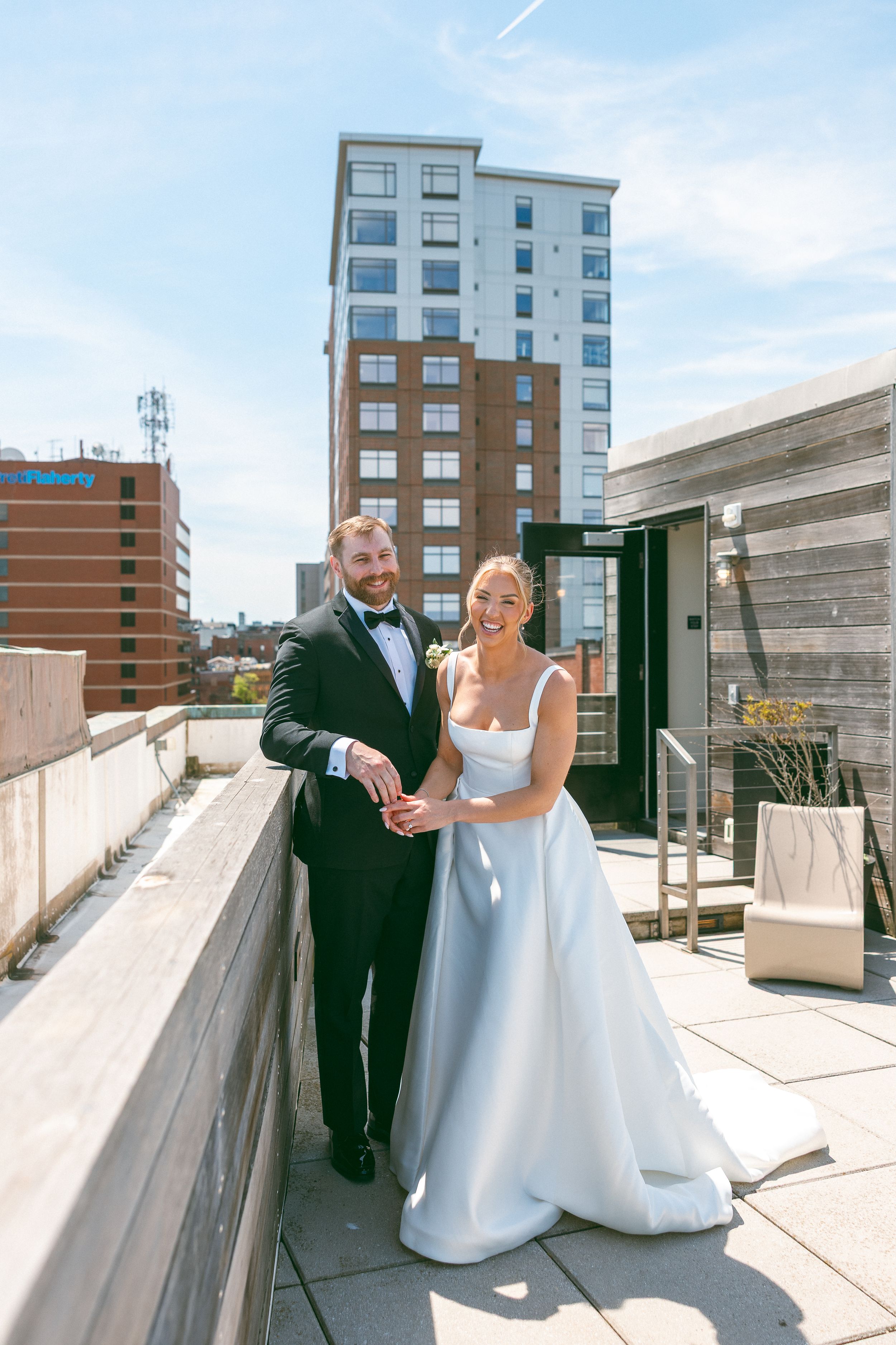 A bride and groom pose together on a rooftop terrace on their wedding day with a city skyline backdrop.