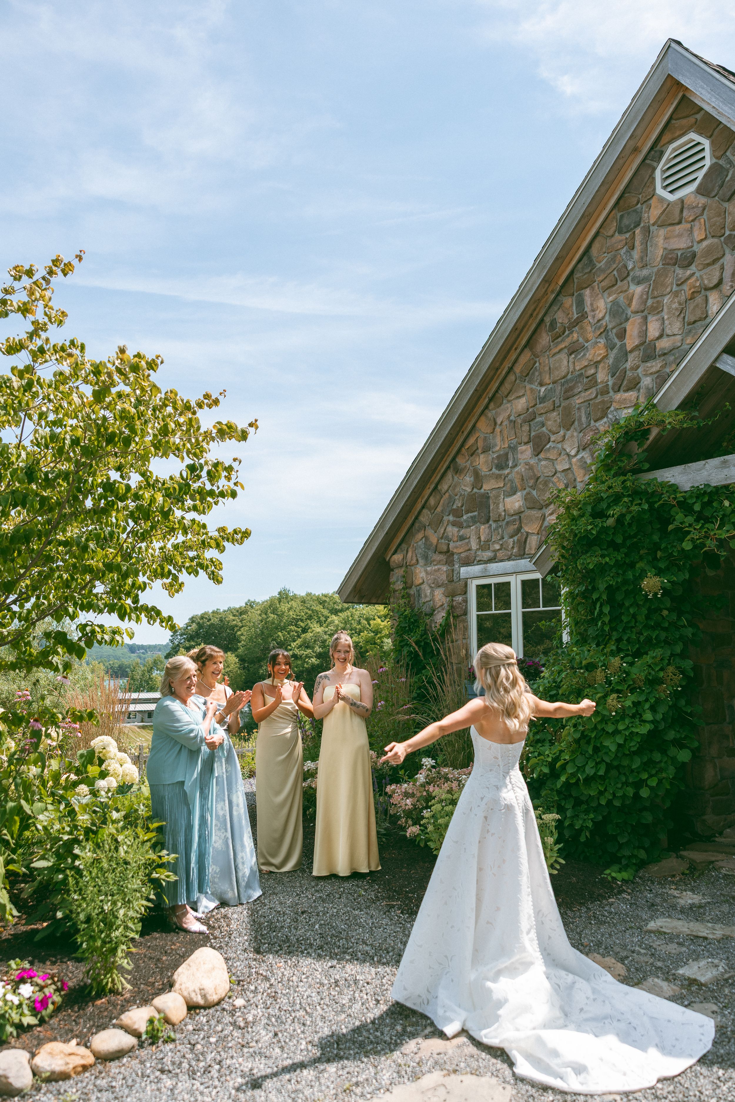 Bride in white gown reveals dress to bridesmaids in sage dresses outside a stone building on a sunny day.