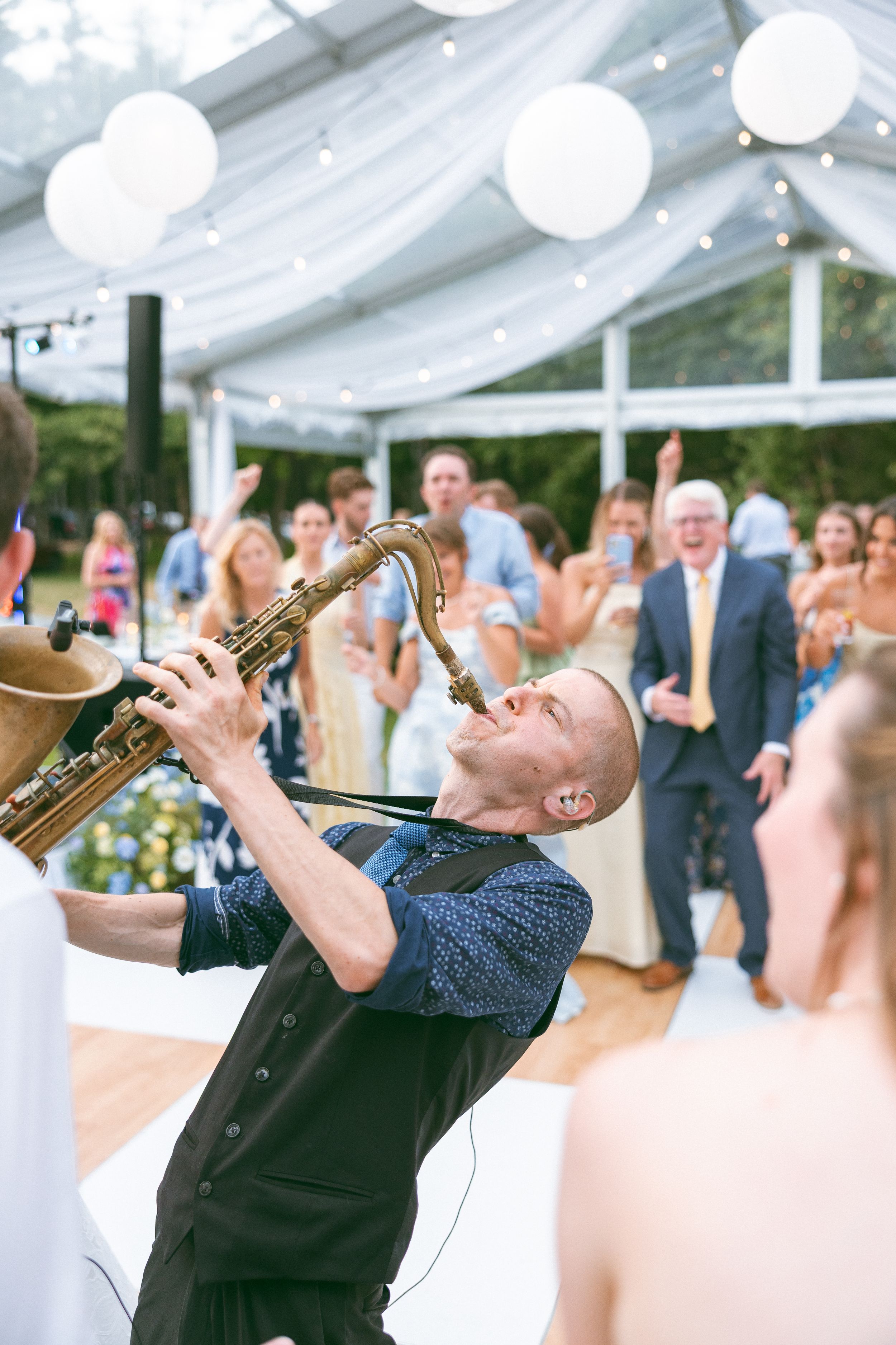 A saxophonist performs energetically at a wedding reception, tilting his head back while guests dance and celebrate in a decorated tent.