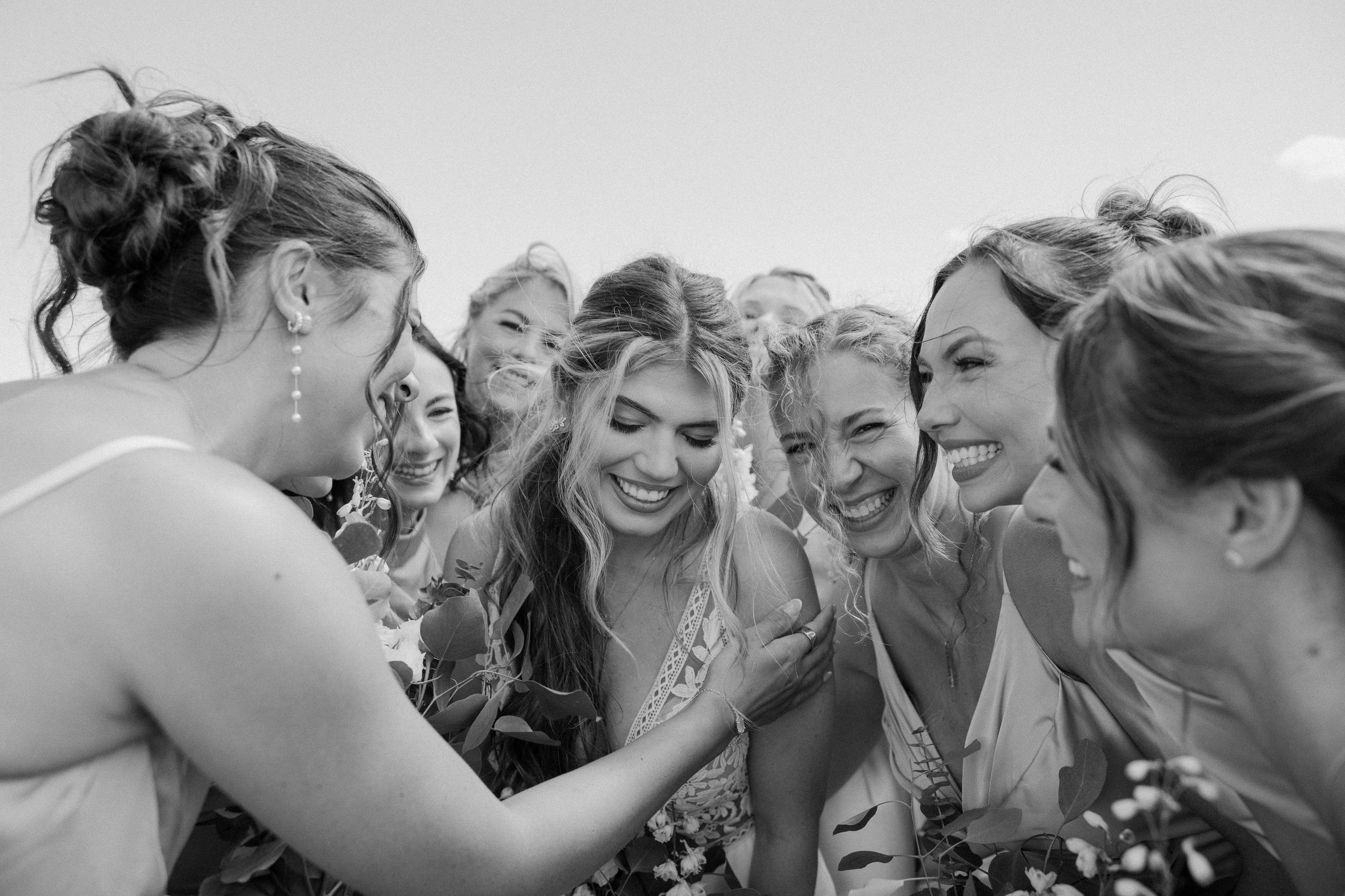 Black and white photo of a bride surrounded by laughing bridesmaids sharing a joyful moment together.