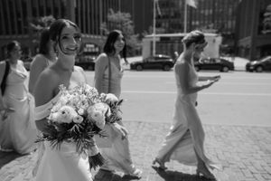 Wedding party members walk along a sunny cobblestone street carrying floral bouquets.