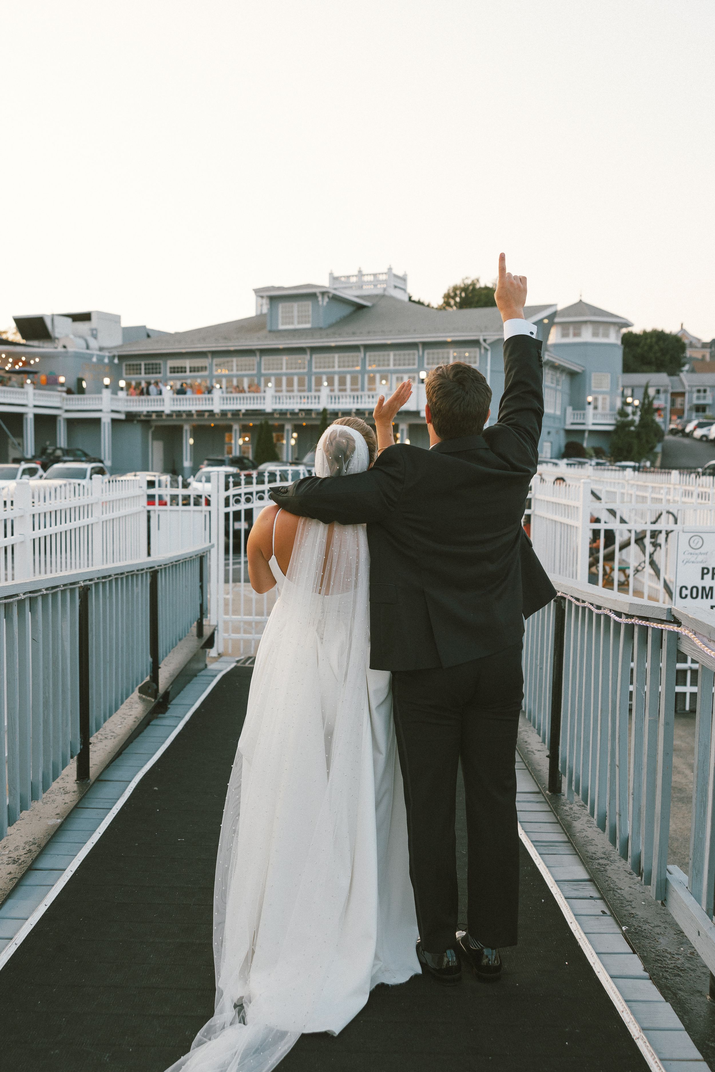 A couple in wedding attire walk along a white railing deck overlooking the water at sunset.