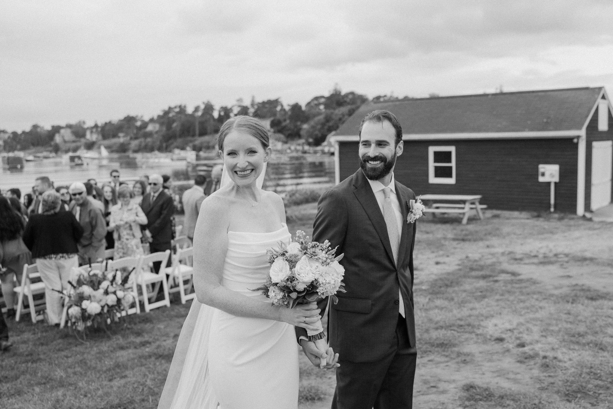 Wedding party processes across a grassy field with rustic buildings in the background.