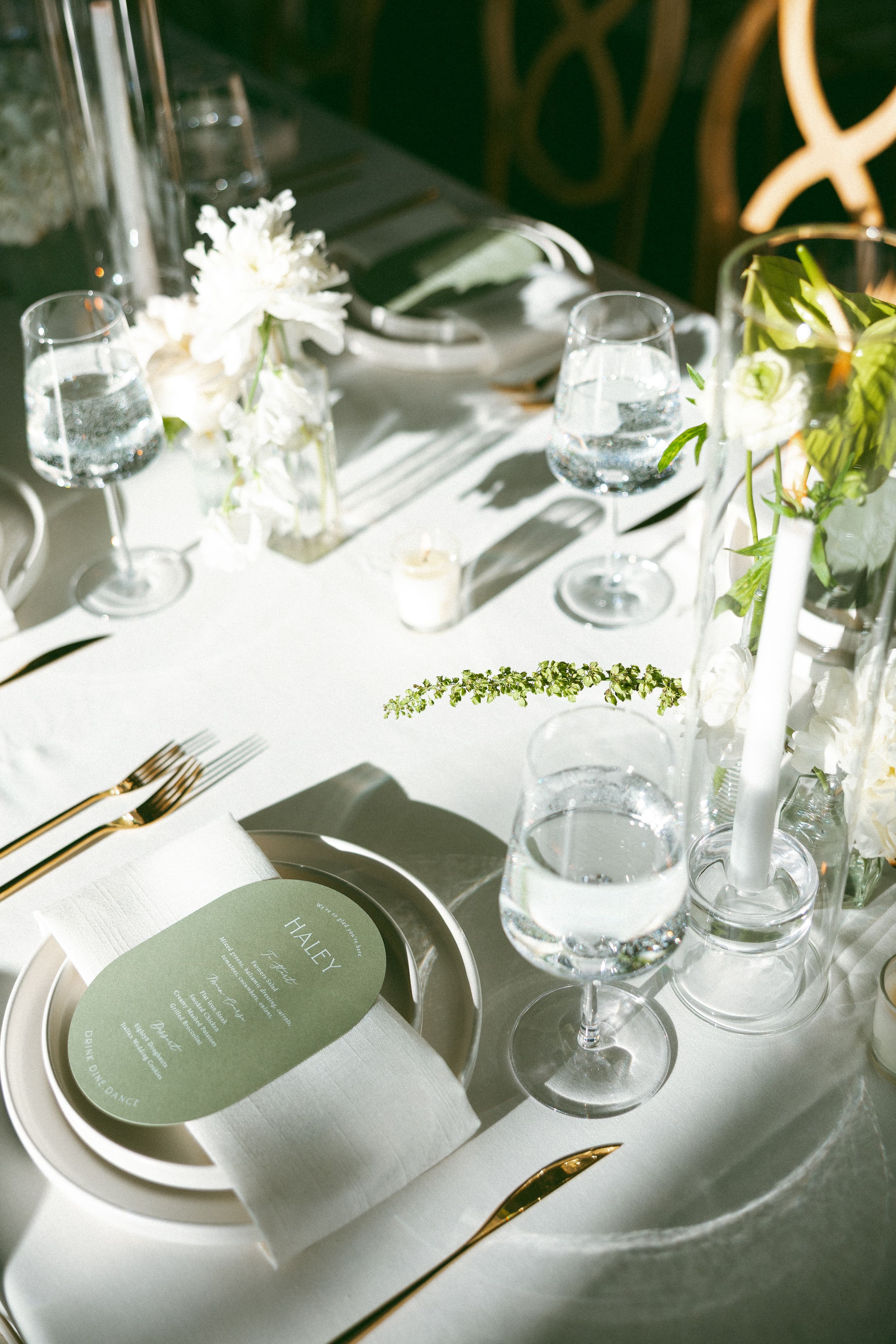 Elegant wedding table setting with white flowers, gold cutlery, sage green menu cards, and crystal glassware on white linen.