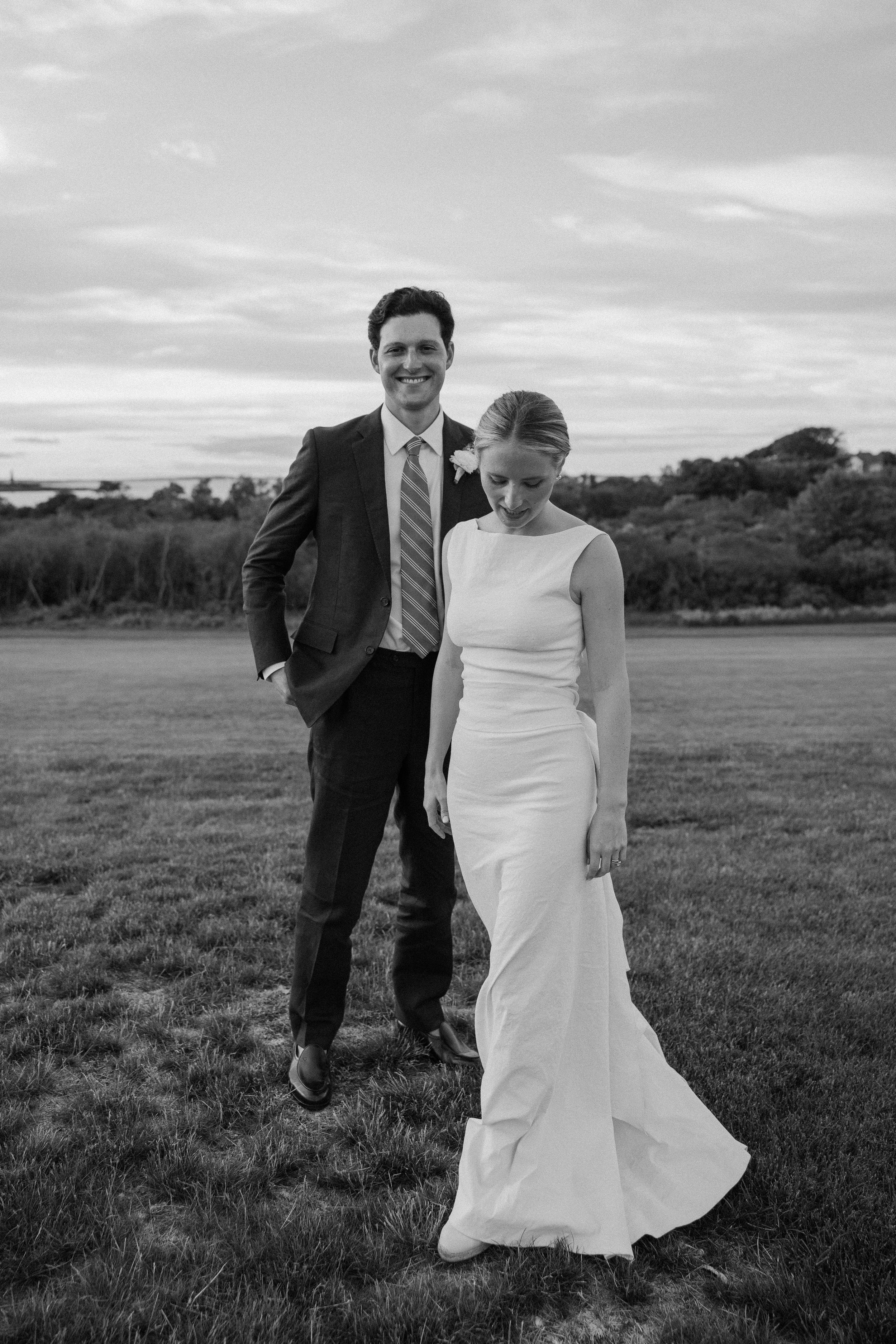 Black and white photo of a smiling groom in a suit and bride in a white gown standing on a grassy field.