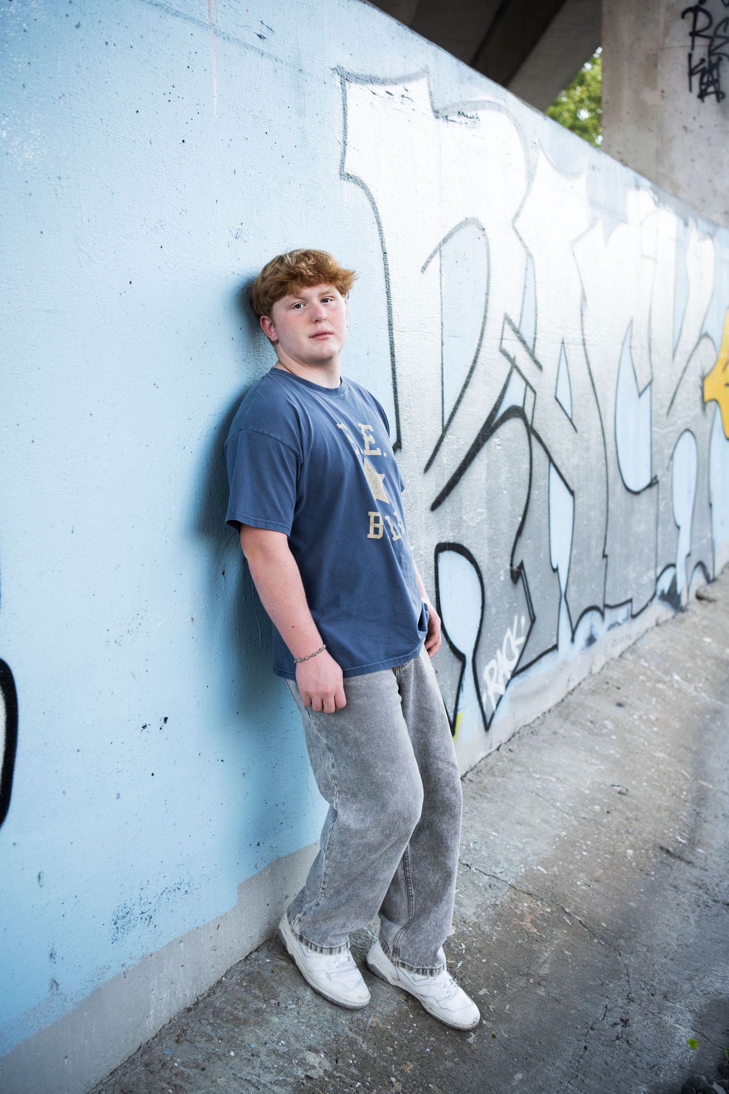 A person in a blue t-shirt and gray pants leans against a wall with graffiti art in an urban setting.
