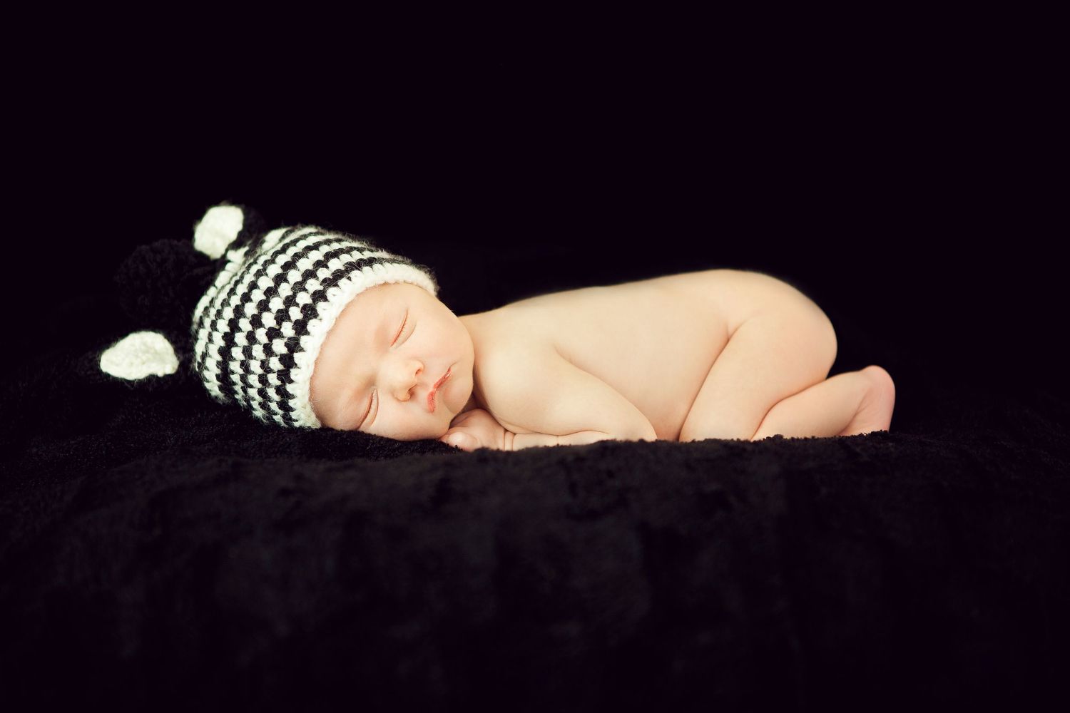 Sleeping newborn baby wearing knitted black and white striped hat curled up on dark background.