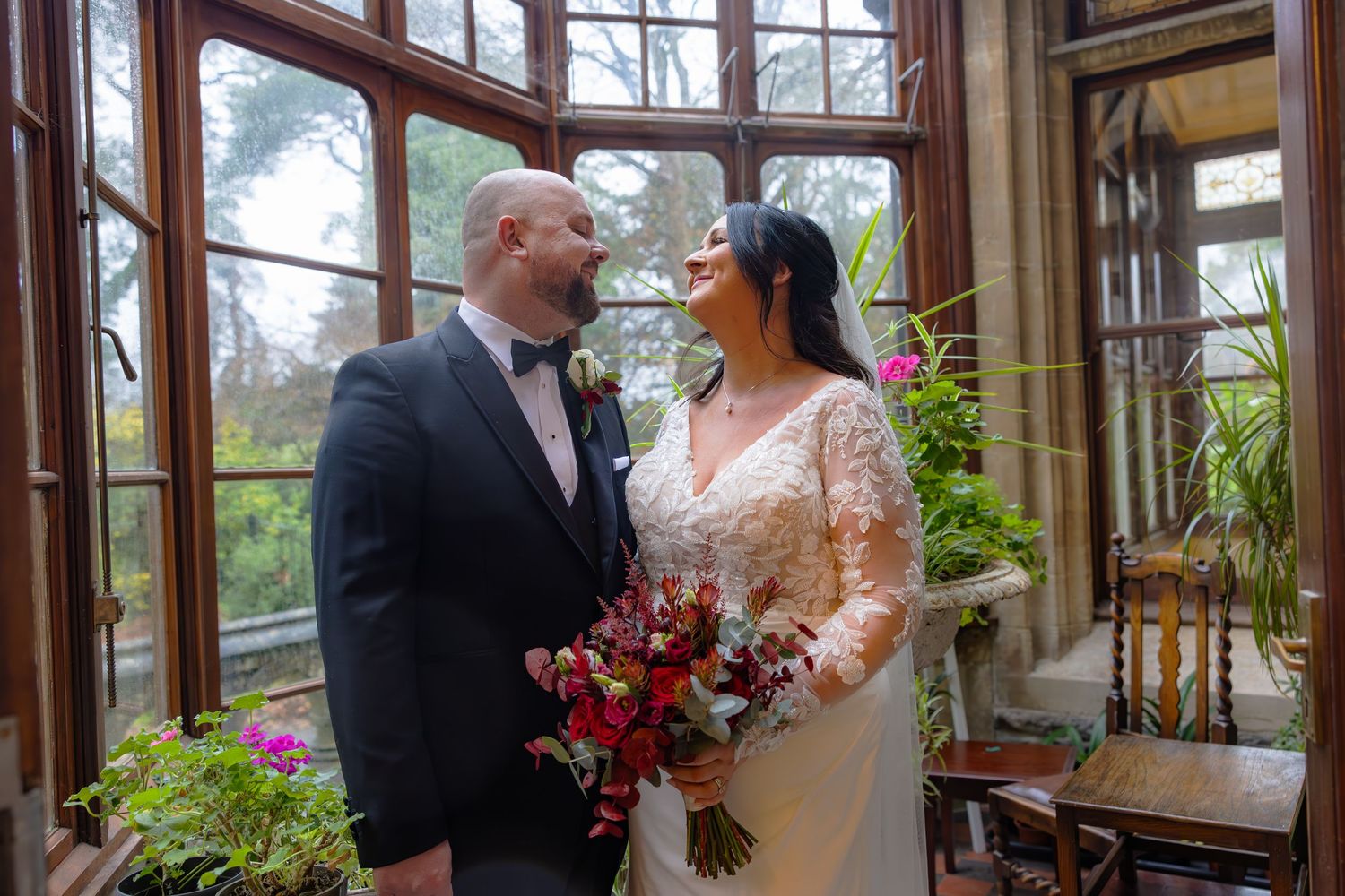 Bride and groom sharing a tender moment in an elegant Victorian greenhouse with colorful bouquet and large windows.