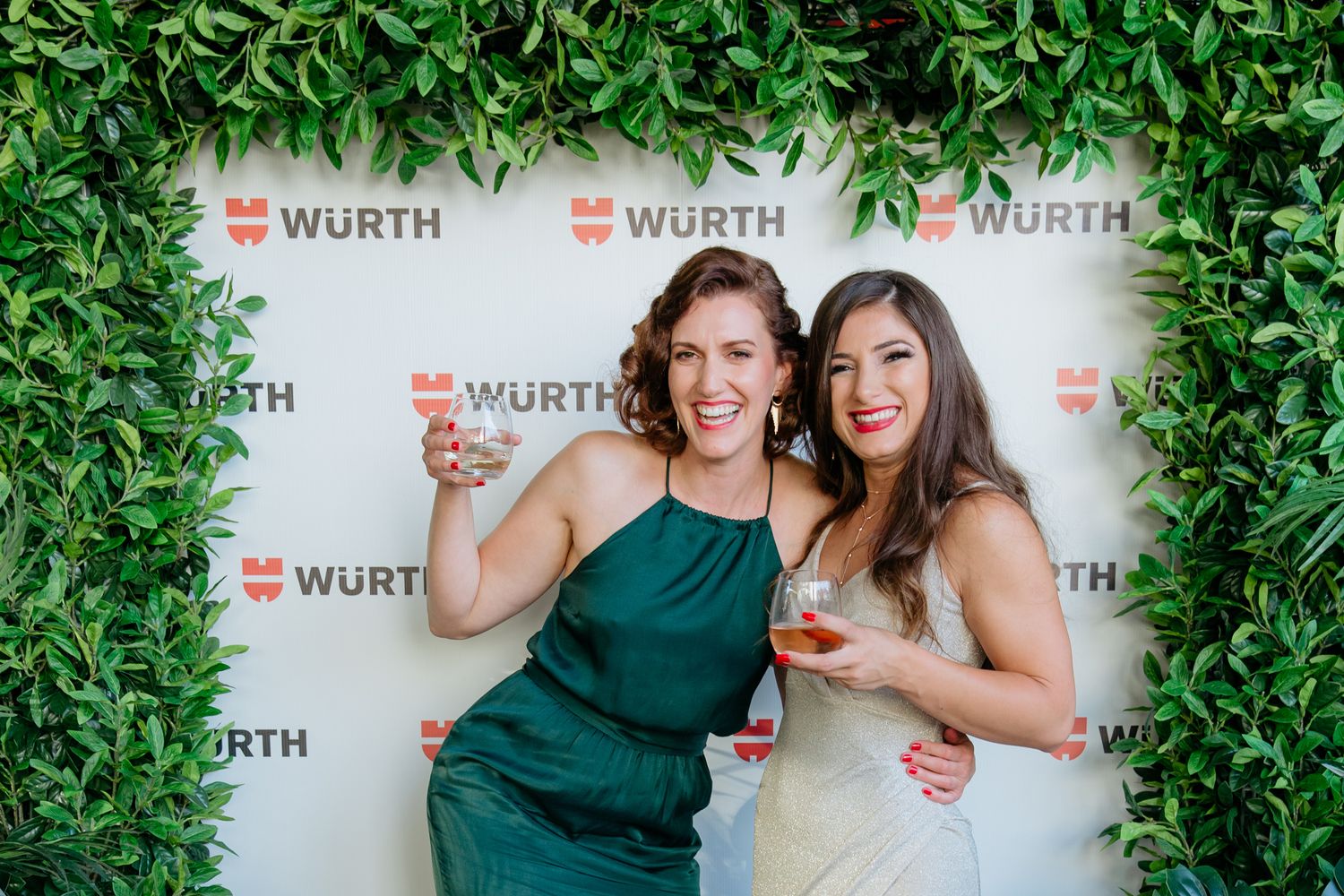 Two women with drinks in hand at a corporate event in front of a media wall with company branding
