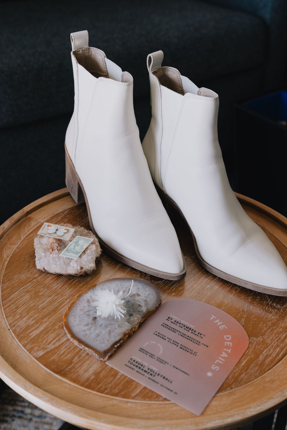 White leather ankle boots displayed on a wooden tray with crystals and wedding stationery details card.