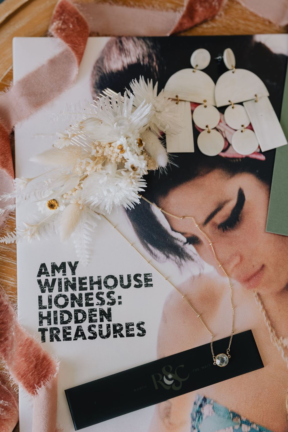 Amy Winehouse Lioness Hidden Treasures album with floral arrangement, earrings, and gold necklace on wooden surface.