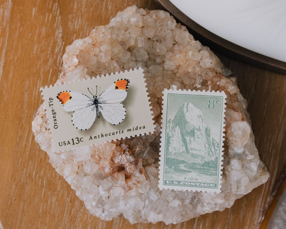 Two vintage US postage stamps displayed on a pink crystal geode, featuring a white butterfly and a scenic mountain landscape.
