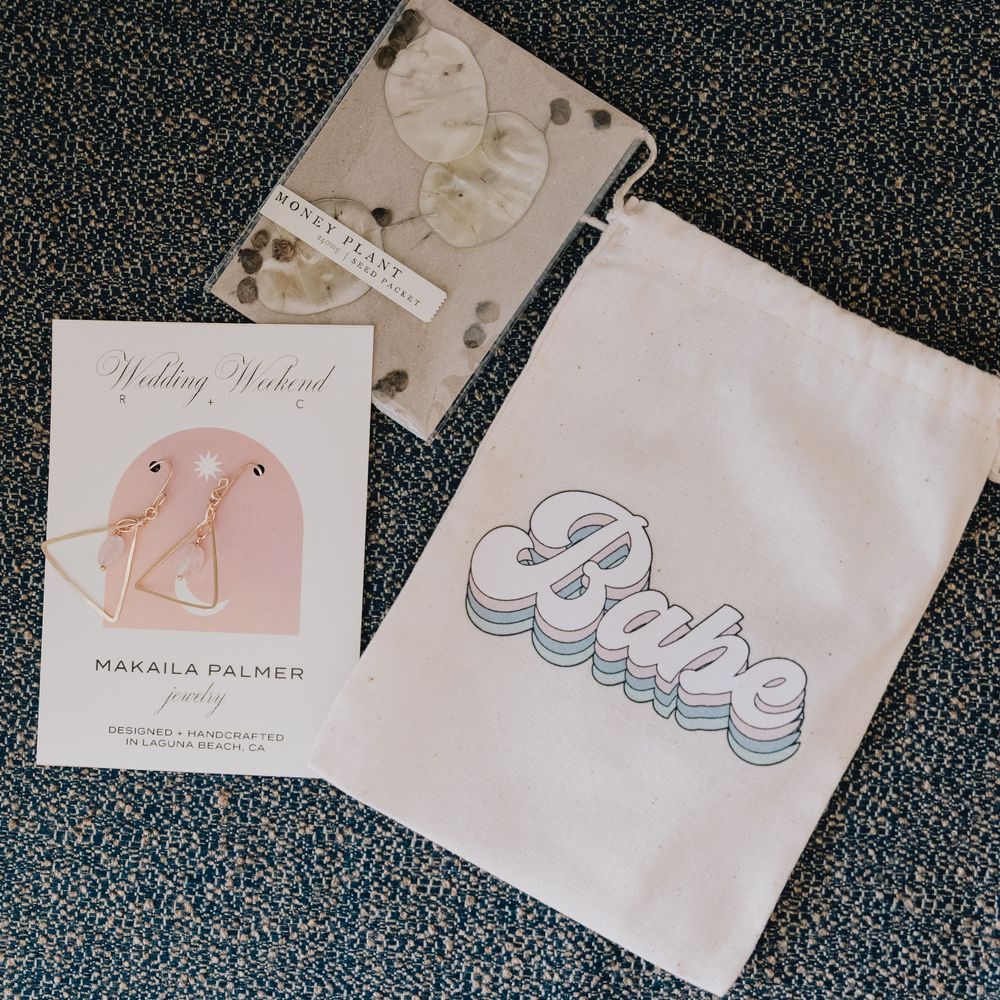 Wedding weekend gift bag with Babe drawstring pouch, hair clip, and floral wax seal on gray carpet.
