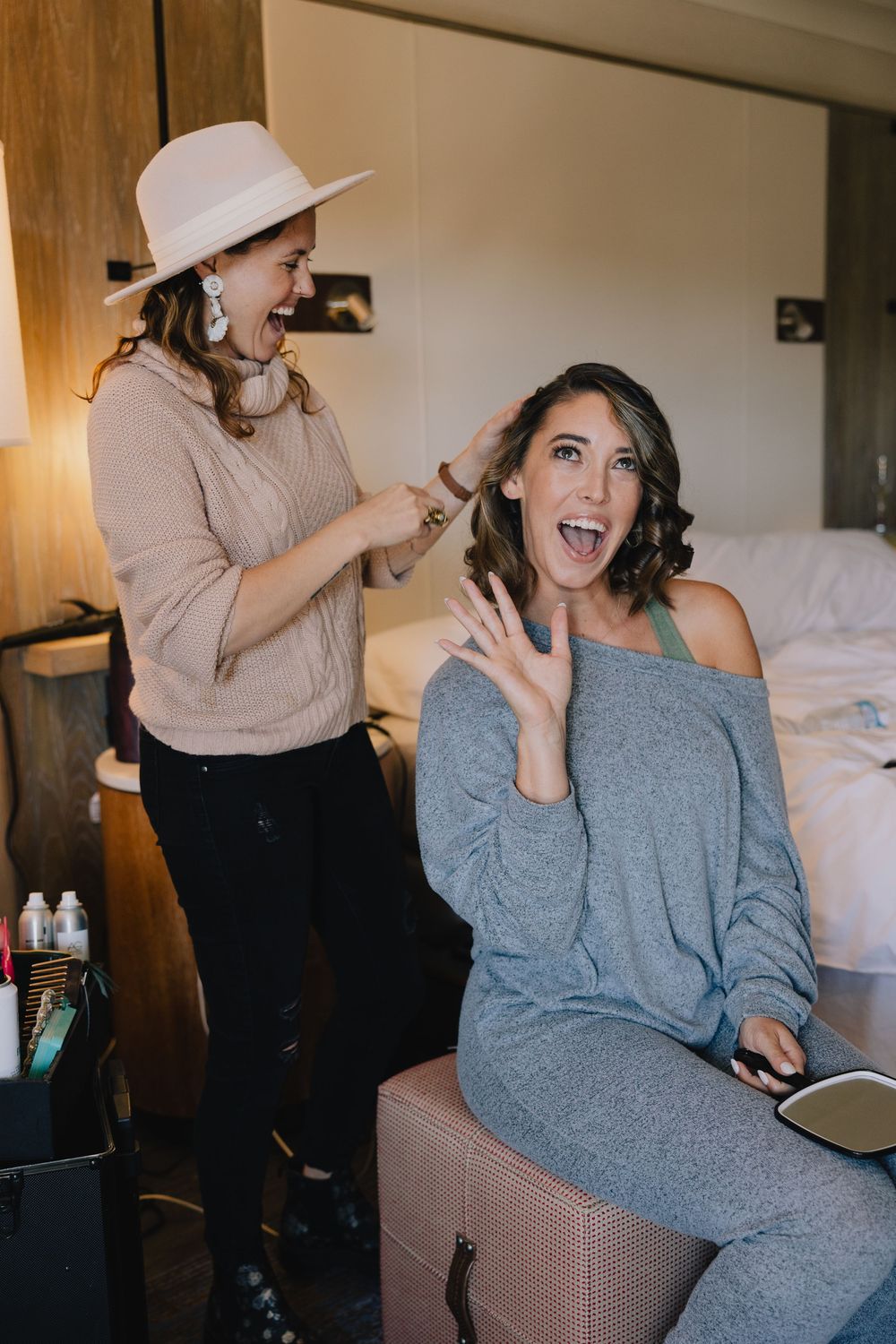 Bride excitedly showing her engagement ring while getting hair styled by bridesmaid in hotel room.