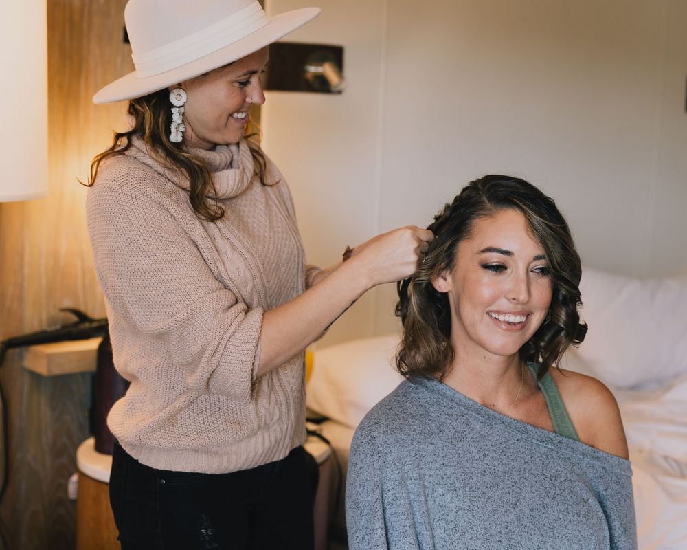 Hairstylist in white hat styling brunette bride's hair in hotel room before wedding ceremony.