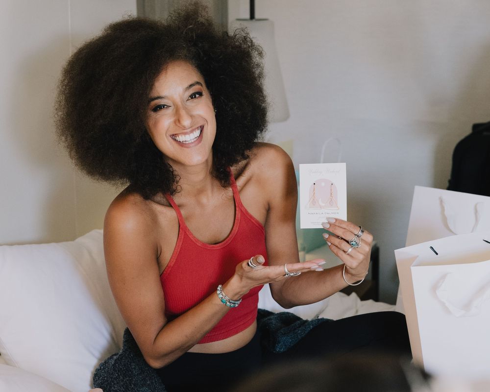 Smiling woman with natural afro hair holding a pink product box while sitting on a white bed.